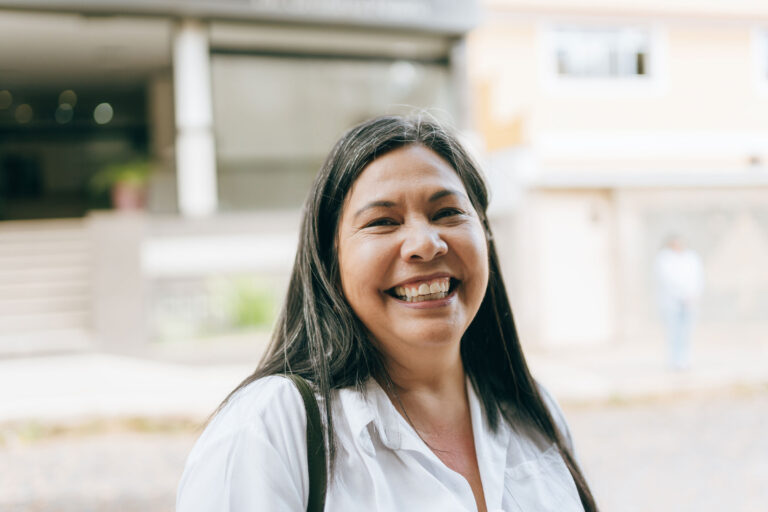 Una mujer sonriendo a la cámara, parada al aire libre frente a un edificio.